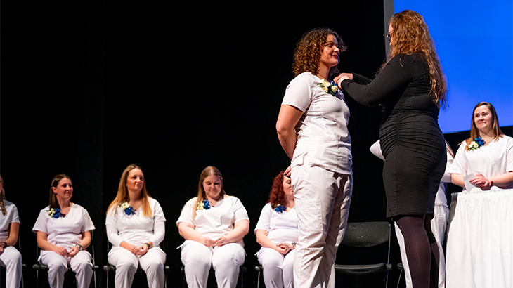 Image: photo of a practical nursing student wearing white scrubs standing on a stage with their instructor who is placing a pin on their blouse, with their other nursing classmates in the background wearing all white scrubs