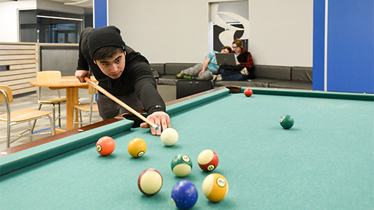 A student playing pool while other students look at a laptop in the background in the lobby area of campus