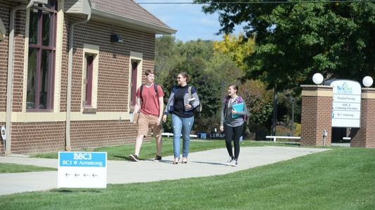 Students walking toward the entrance of BC3 Armstrong with a lawn sign in the foreground that has arrows pointing to the left and the text "BC3 @ Armstrong" in blue letters