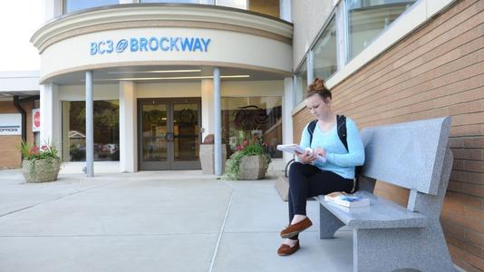A female student with a light blue shirt sits on a stone bench reading a textbook directly outside of the entrance to the BC2 @ Brockway building