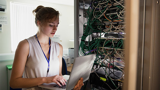 Cybersecurity employee working on laptop in a server room with wires and a server in the background