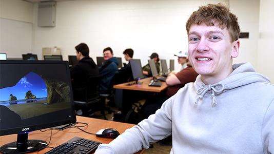 Image: portrait photo of a male student sitting in a computer lab with a desktop computer to his left