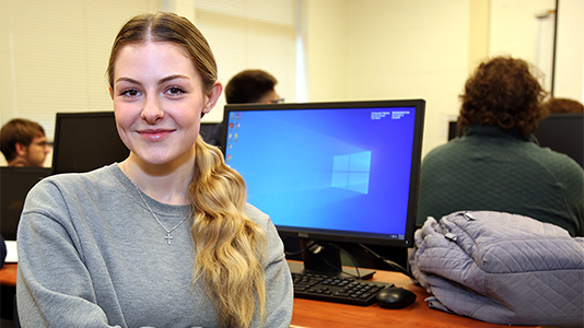 Image: portrait photo of a female student smiling with a desktop computer behind her on a desk