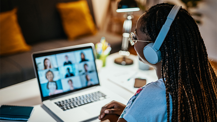 Image: a woman wearing white headphones while looking at a laptop screen with a video conference call of different people's faces