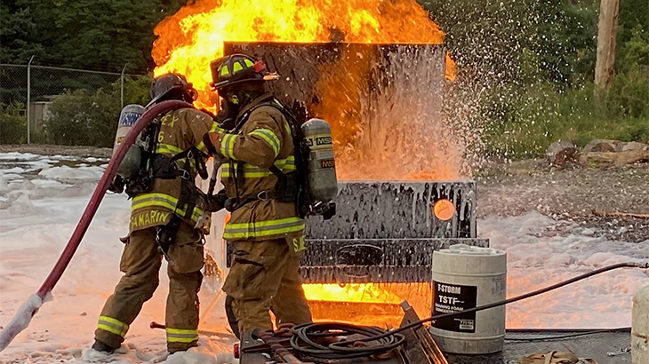 Image: photo of a simulated fire with two students dressed in firefighting gear putting out a fire using foam, with orange flames in the background