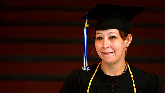 Image: a female graduate wearing a black cap and gown and gold cords