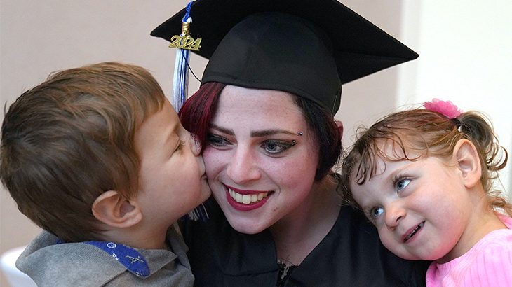 Image: a photo of a smiling woman wearing a black cap and gown hugging two children